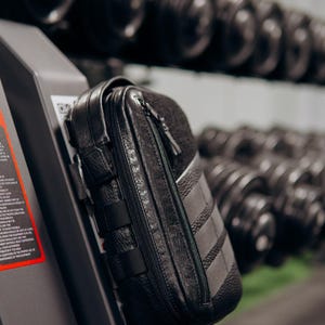 Op de afbeelding: Zwarte leren etui met ritssluiting en meerdere bandjes. De etui is bevestigd aan een fitnessapparaat, met een rek met dumbbells op de achtergrond. De etui heeft een textuur en een contrasterend grijs paneel.