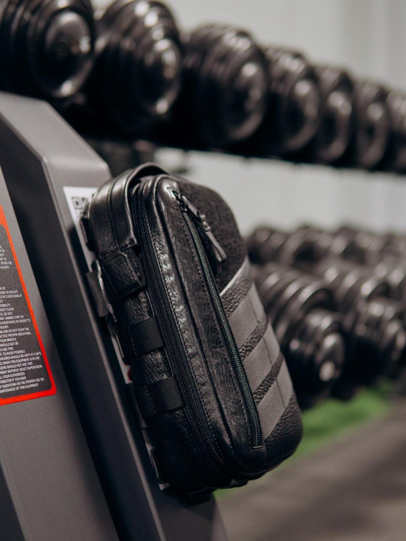 Op de afbeelding: Zwarte leren etui met ritssluiting en meerdere bandjes. De etui is bevestigd aan een fitnessapparaat, met een rek met dumbbells op de achtergrond. De etui heeft een textuur en een contrasterend grijs paneel.