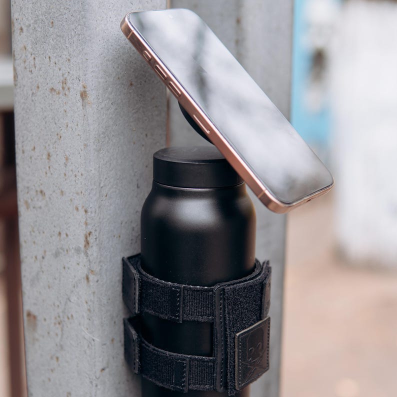 Op de afbeelding: Een zwarte ge&iuml;soleerde waterfles met een zwarte klittenbandriem en een smartphone bevestigd. De telefoon heeft een goudkleurig frame. De fles is op een metalen paal gemonteerd. De achtergrond is wazig.