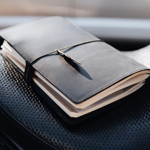 Op de afbeelding: Een donkergrijze lederen notitieboekhoes met een gladde, matte afwerking en zichtbare stiksels langs de randen. Het notitieboek wordt vastgehouden door een dun zwart elastisch koord dat om het midden is gewikkeld, met een kleine metalen kruisvormige bedel.