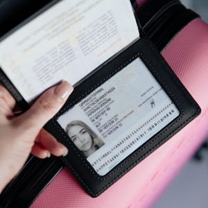 May include: Black leather passport holder with a clear window displaying a passport and identification card. The ID card shows a photo and personal details. The holder is open, revealing the passport pages. The background is a pink suitcase.