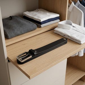 May include: A black leather belt with a silver-toned metal buckle and a single loop, folded into a compact shape. The belt features a smooth finish and a metallic stud near the end, resting on a light-colored wooden shelf in a closet.