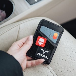 May include: Black leather wallet with a skull patch and the word "RIDL" on a patch. A credit card is visible in a pocket. The wallet is held in a hand.