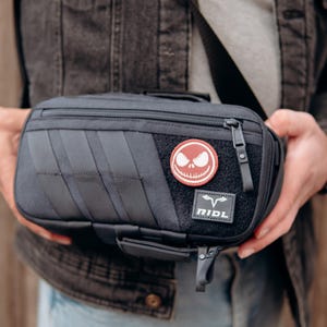 Op de afbeelding: Zwarte rechthoekige tas met ritssluiting en een klein handvat. De tas heeft een patch met een Jack Skellington-ontwerp en een logo met de tekst "RIDL". De tas heeft een riem en wordt door een persoon vastgehouden.
