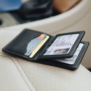 Può includere: Portafoglio nero in pelle a due ante con carte di credito e d'identità visibili. Il portafoglio è aperto, mostrando i compartimenti interni e le carte. Cuciture bianche. Su una superficie chiara.