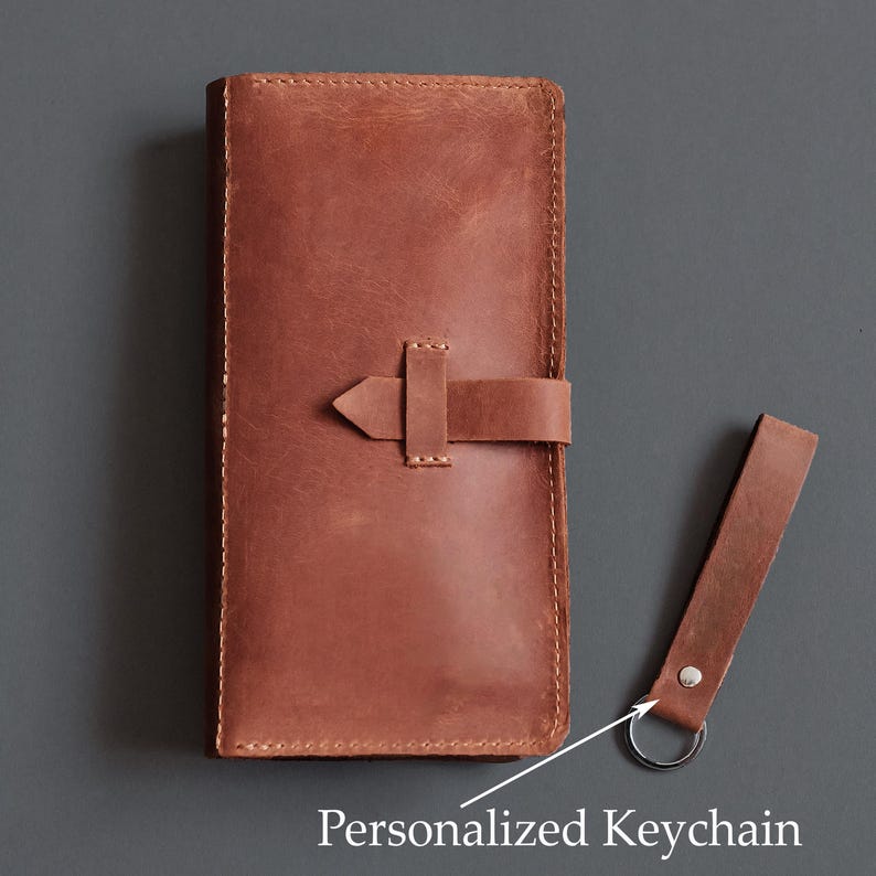 May include: A rectangular brown leather wallet with visible stitching and a horizontal strap closure, accompanied by a matching brown leather keychain featuring a silver metal ring and the text Personalized Keychain.