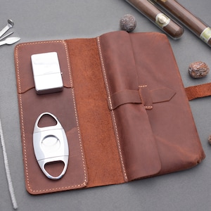 May include: A brown leather cigar case featuring a fold-over flap with a strap closure. The interior includes dedicated pockets holding a silver metal lighter and a silver cigar cutter. The case is constructed with visible stitching along the edges.