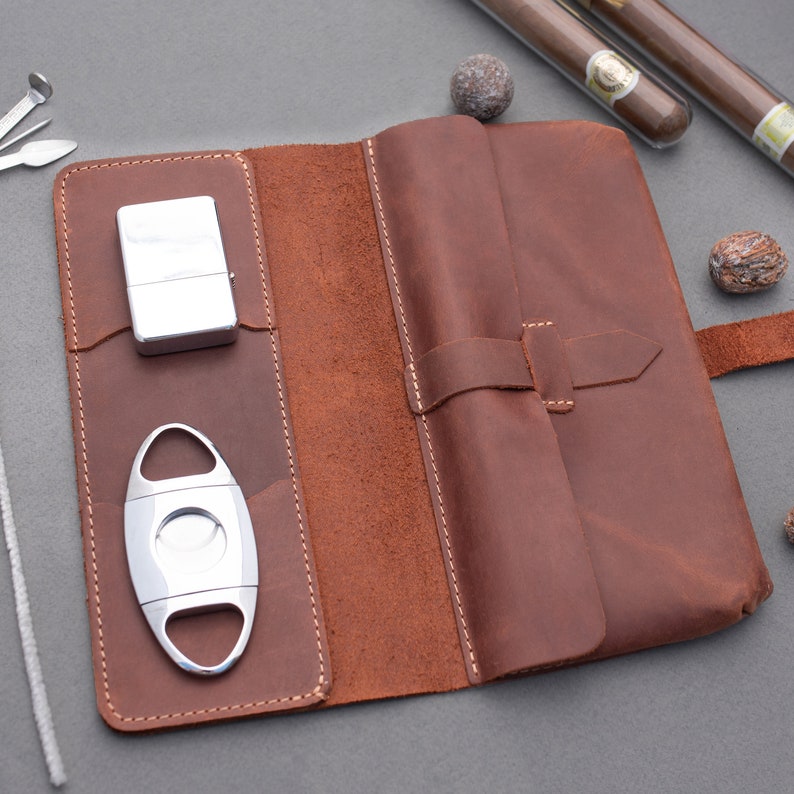 May include: A brown leather cigar case featuring a fold-over flap with a strap closure. The interior includes dedicated pockets holding a silver metal lighter and a silver cigar cutter. The case is constructed with visible stitching along the edges.