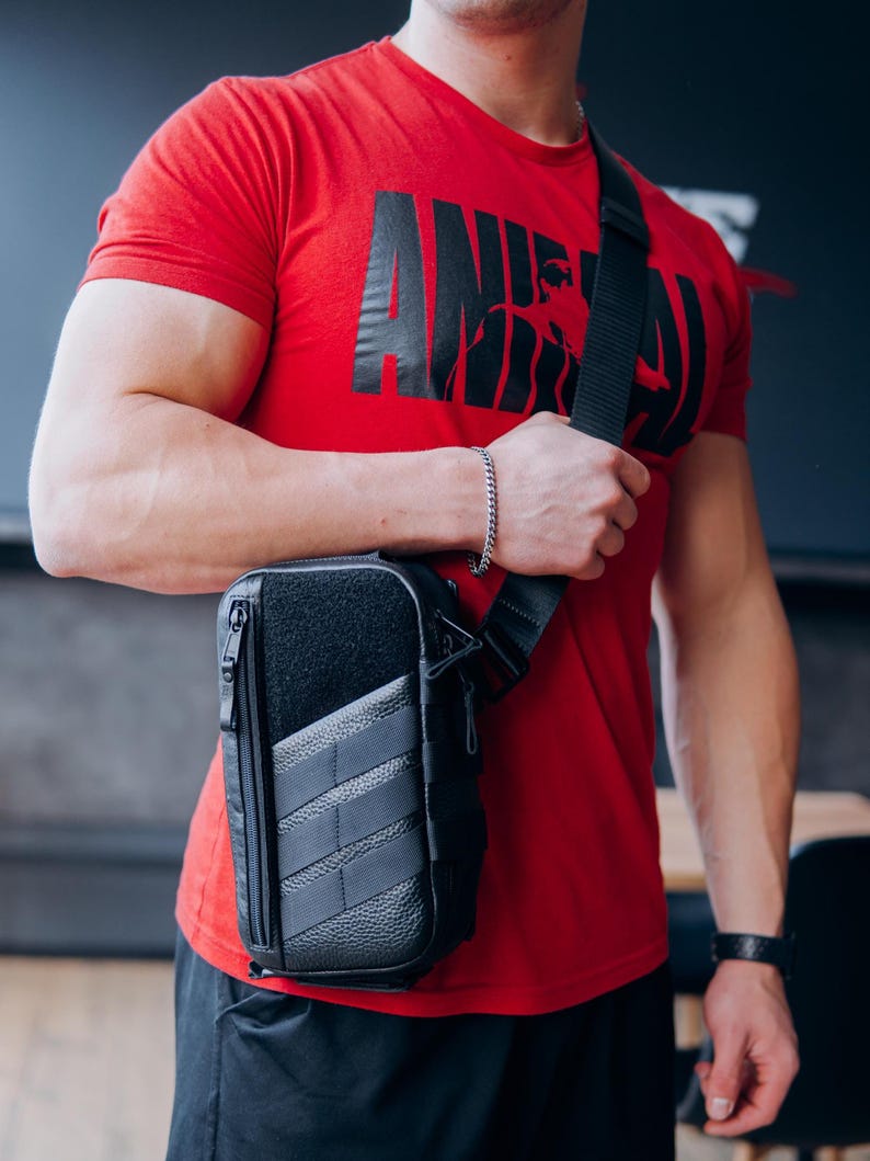 Op de afbeelding: Zwarte crossbodytas met een getextureerd voorpaneel en een vak met ritssluiting. De tas heeft een zwarte, verstelbare schouderriem. De persoon op de foto draagt een rood t-shirt met de zwarte tekst "ANIMAL".