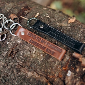 May include: Two leather keychains with metal rings and the text "KEYS TO MY FUCKING AWESOME BIKE" embossed on them. One keychain is brown, the other is black. The keychains are resting on a textured, brown surface.