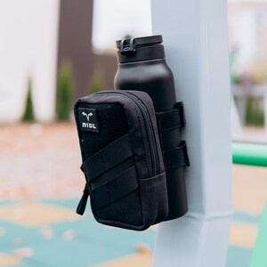 Op de afbeelding: Zwarte ge&iuml;soleerde waterfles met een zwart zakje. Het zakje heeft een rits en de tekst "RIDL" op een patch. De fles is vastgezet met zwarte bandjes. De items zijn gemonteerd op een witte paal.