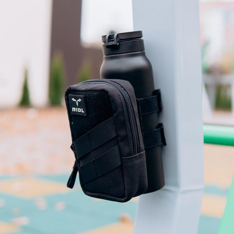 Op de afbeelding: Zwarte ge&iuml;soleerde waterfles met een zwart zakje. Het zakje heeft een rits en de tekst "RIDL" op een patch. De fles is vastgezet met zwarte bandjes. De items zijn gemonteerd op een witte paal.
