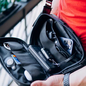 Op de afbeelding: Zwarte crossbodytas met meerdere open vakken. Zichtbare items zijn een smartphone, sleutels, een draagbare oplader, een creditcard en witte oortelefoons. De tas heeft een zwarte riem en een zwarte ritssluiting.