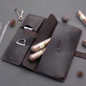 May include: A dark brown leather cigar case featuring multiple interior pockets, containing two cigars in clear plastic tubes, a silver metal cigar cutter, and a multi-tool accessory. The case has visible stitching along the edges and a flap closure.
