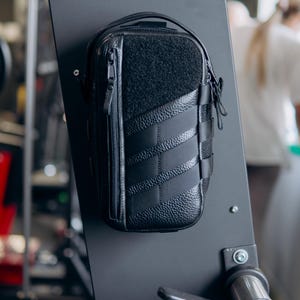 Op de afbeelding: Zwarte rechthoekige etui met een getextureerd voorpaneel en een ritssluiting. De etui heeft een lusgreep en is bevestigd aan een zwart metalen oppervlak. Het ontwerp omvat gestikte diagonale lijnen en een klittenband.