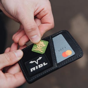 May include: A black leather wallet with a pirate-themed patch being inserted. The patch is square, with a green background and a skull and crossbones design. A credit card is visible in a slot. The wallet also has a white logo that says "RIDL".