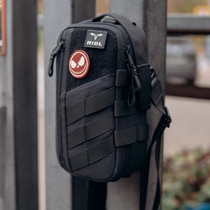 Op de afbeelding: Zwarte tactische schoudertas met meerdere banden en een hoofdvak met ritssluiting. Voorzien van een patch met het woord "RIDL" en een ronde patch met een Jack Skellington-ontwerp. De tas hangt aan een metalen paal.