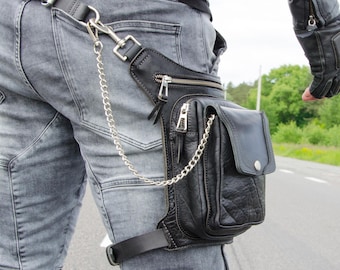 May include: A black leather drop leg bag featuring multiple zippered compartments, a front flap pocket with a snap closure, and a silver-toned metal chain attachment. The bag is secured to the thigh with an adjustable strap and to the waist belt.
