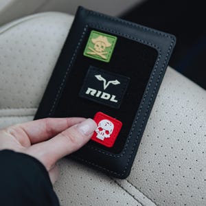 May include: Black leather wallet with a black fabric patch. The wallet has three patches: a green square with a skull and crossbones, a black patch with the word "RIDL", and a red square with a white skull.