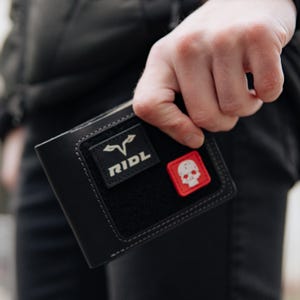 May include: Black leather wallet with two patches. One patch is black with the word "RIDL" and a stylized design. The other patch is red with a white skull. The wallet is held in a hand.
