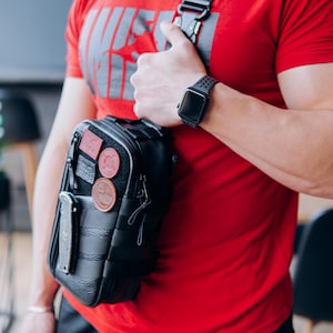 Op de afbeelding: Zwarte crossbodytas met meerdere patches en een mes. De tas heeft een zwarte band en wordt over de schouder gedragen. De persoon draagt een rood t-shirt en een zwarte smartwatch.