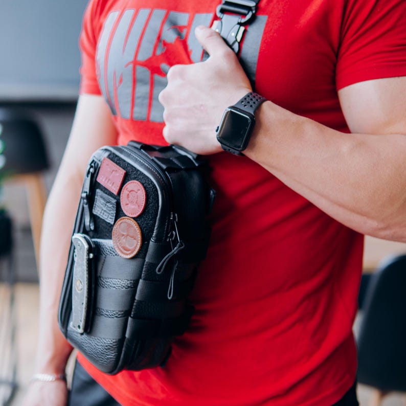 Op de afbeelding: Zwarte crossbodytas met meerdere patches en een mes. De tas heeft een zwarte band en wordt over de schouder gedragen. De persoon draagt een rood t-shirt en een zwarte smartwatch.