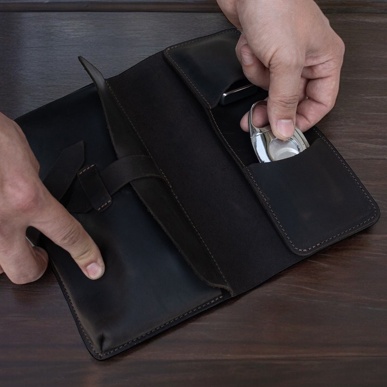 May include: A dark brown leather case featuring multiple interior pockets and a strap closure. The case contains a metallic silver cigar cutter in one of the slots. The leather has visible stitching along the edges and a smooth, matte finish.