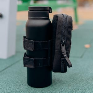 Op de afbeelding: Een matzwarte ge&iuml;soleerde waterfles met een bijpassend zwart etui. De fles is vastgezet met twee zwarte bandjes. Het etui heeft een ritssluiting en een kleine lus. De items staan op een groene ondergrond.