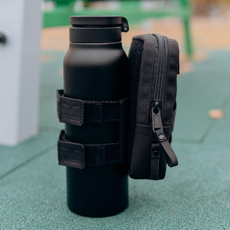 Op de afbeelding: Een matzwarte ge&iuml;soleerde waterfles met een bijpassend zwart etui. De fles is vastgezet met twee zwarte bandjes. Het etui heeft een ritssluiting en een kleine lus. De items staan op een groene ondergrond.