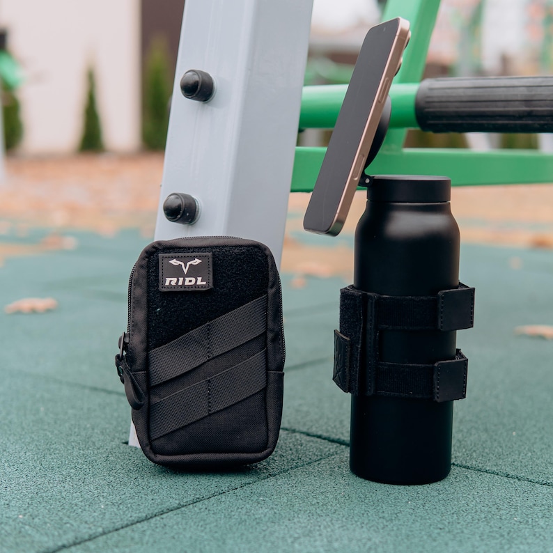 Op de afbeelding: Zwarte ge&iuml;soleerde waterfles met een telefoon bevestigd, vastgehouden door een zwarte band. Een zwart etui met de tekst "RIDL" staat ook in de afbeelding. De items staan op een groene ondergrond.