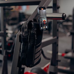 Op de afbeelding: Zwarte leren telefoonhoes met een ritsvak en een klittenband patch, gemonteerd op een metalen gymrek. Een smartphone is bevestigd, met een video-opname-interface. De sportschoolomgeving omvat trainingsapparatuur.