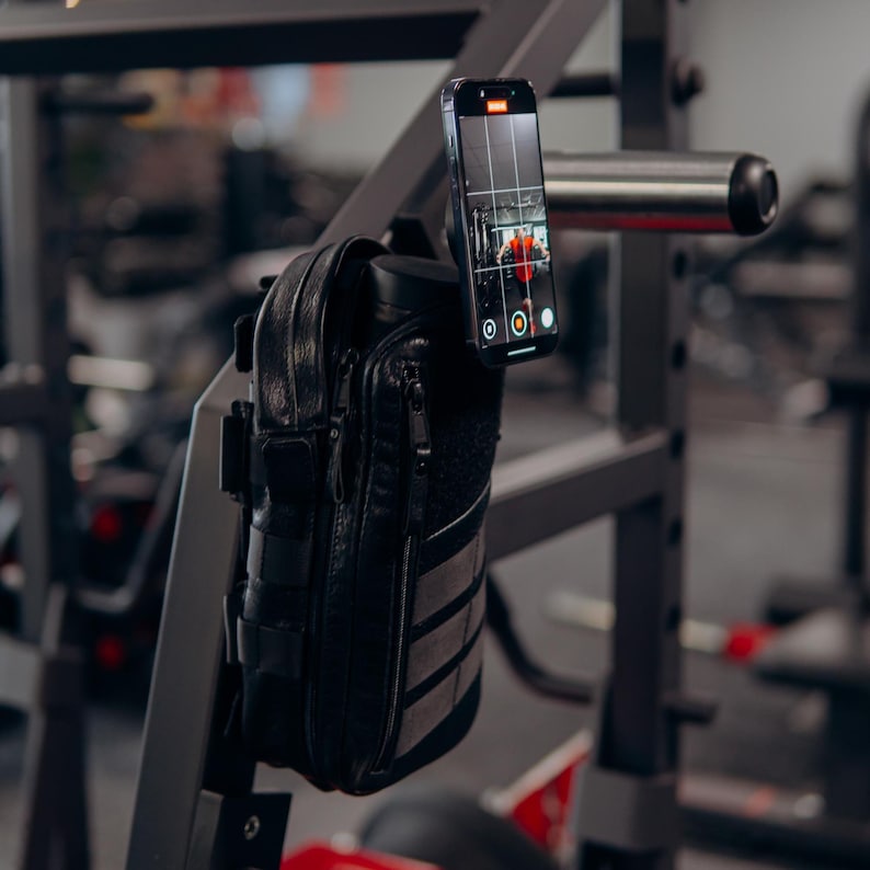 Op de afbeelding: Zwarte leren telefoonhoes met een ritsvak en een klittenband patch, gemonteerd op een metalen gymrek. Een smartphone is bevestigd, met een video-opname-interface. De sportschoolomgeving omvat trainingsapparatuur.