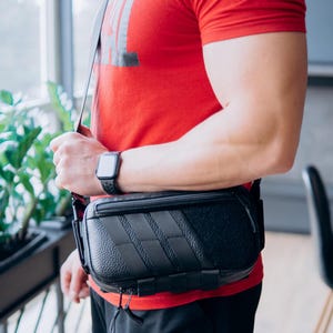 Op de afbeelding: Zwarte crossbodytas met een getextureerd voorpaneel en een ritssluiting aan de bovenkant. De tas heeft een schouderriem. De persoon die de tas vasthoudt, draagt een rood t-shirt en een zwart horloge.