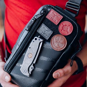 Op de afbeelding: Zwarte crossbodytas met meerdere leren patches en een zilveren vouwmes. De tas heeft een textuurontwerp en een zwarte riem. De patches zijn in verschillende kleuren, waaronder rood en bruin. Het mes is zilver en grijs.