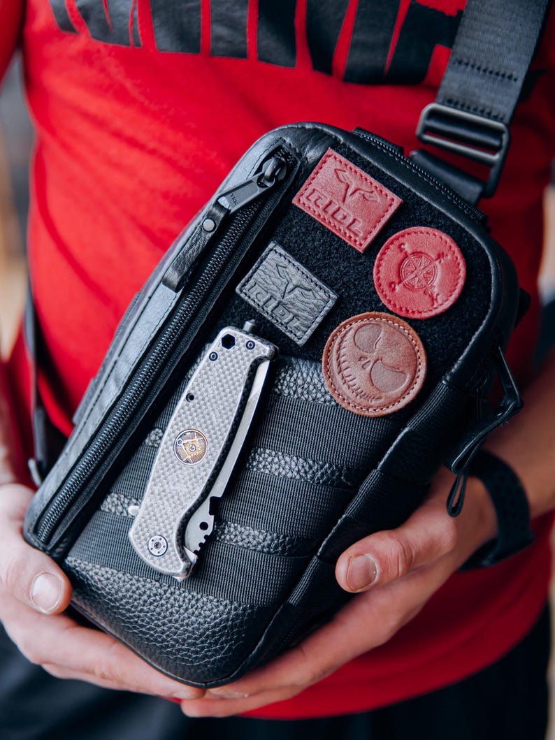 Op de afbeelding: Zwarte crossbodytas met meerdere leren patches en een zilveren vouwmes. De tas heeft een textuurontwerp en een zwarte riem. De patches zijn in verschillende kleuren, waaronder rood en bruin. Het mes is zilver en grijs.