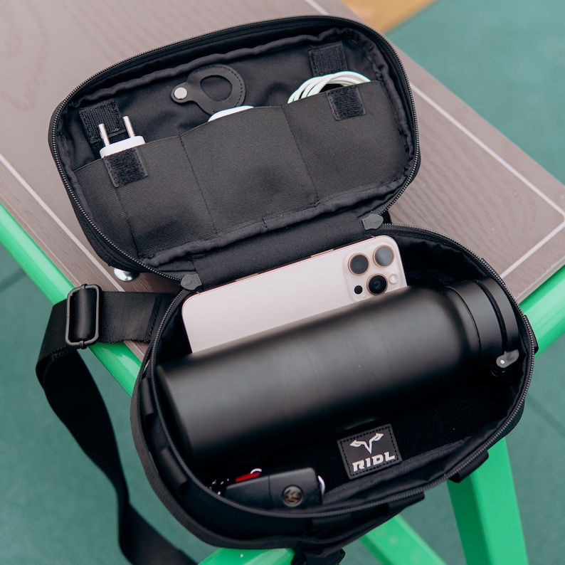 Op de afbeelding: Zwarte crossbodytas met een open bovenkant die een smartphone, een zwarte waterfles en diverse accessoires laat zien. De tas heeft een schouderriem en een klein logopatch. De binnenkant heeft meerdere vakken.