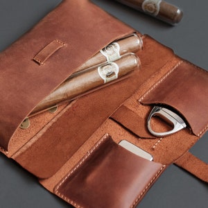 May include: A brown leather cigar case featuring two cigars in clear glass tubes, a silver metal cigar cutter, and a small silver metal lighter. The case has dedicated interior pockets for each item and is constructed with visible stitching.