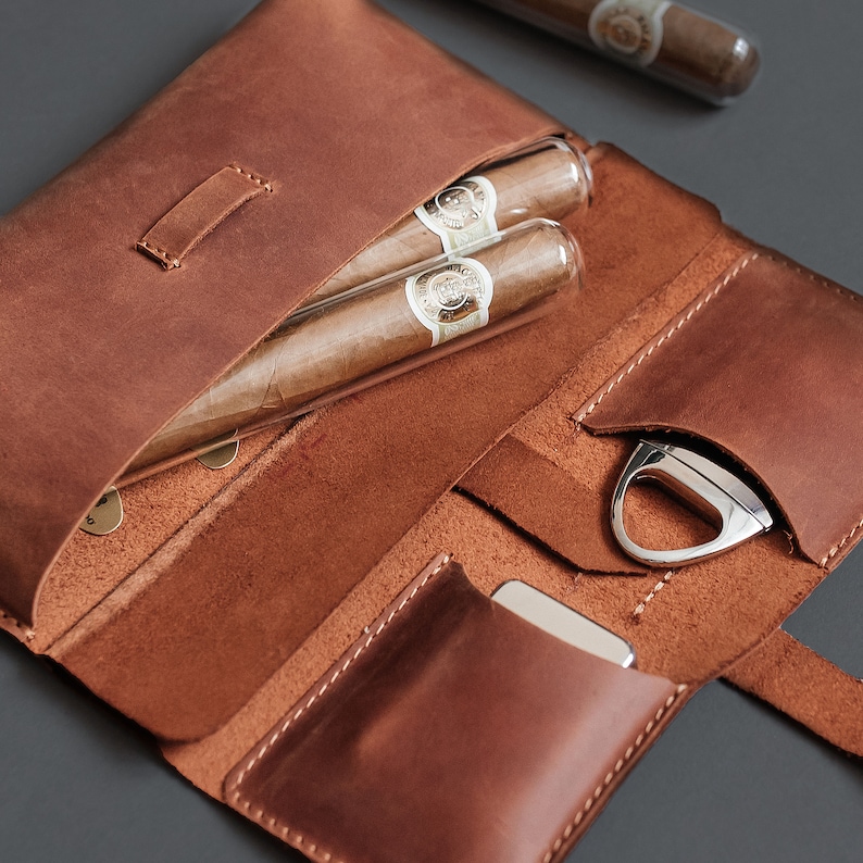 May include: A brown leather cigar case featuring two cigars in clear glass tubes, a silver metal cigar cutter, and a small silver metal lighter. The case has dedicated interior pockets for each item and is constructed with visible stitching.