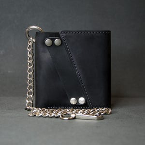 May include: Black leather wallet with a silver chain. The wallet features a flap closure secured by two silver rivets. A silver chain is attached to the wallet with a key ring clasp. The wallet is rectangular and has a minimalist design.