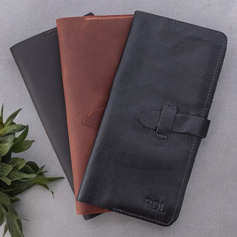 May include: Three rectangular leather wallets in black, brown, and dark gray, arranged in a fan shape. The black wallet features a horizontal strap closure and the embossed letters RDL in the bottom corner, with visible stitching along the edges.