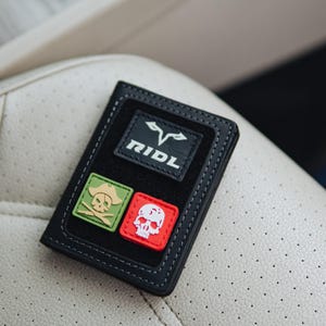 May include: Black wallet with white stitching and three patches. The top patch has the word "RIDL" and a logo. The other patches are a green skull and crossbones and a red skull. The wallet is on a cream-coloured surface.