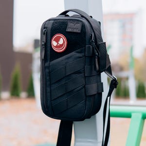 Op de afbeelding: Zwarte tactische schoudertas met meerdere bandjes en een patch met een rood-wit schedelontwerp. De tas heeft een handvat aan de bovenkant, een hoofdvak met ritssluiting en een schouderriem. De tas is ontworpen voor het dragen van benodigdheden.