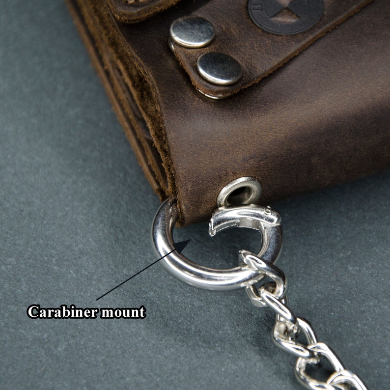 May include: A brown leather wallet with a silver metal carabiner mount and chain. The wallet has a worn look and is made of high-quality leather.