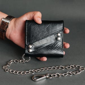 May include: A black leather bifold wallet featuring a textured surface, a diagonal strap detail secured with four silver-toned metal rivets, and a long silver-toned metal chain attached to a circular ring on the bottom corner of the wallet.
