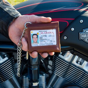 May include: A brown leather wallet with a chain attached. The wallet has a clear plastic window displaying an Alabama driver's license with the name Connor Sample. The wallet is held in a hand in front of a black motorcycle.