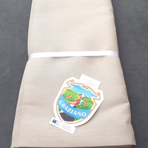 May include: Rolled beige linen fabric, tied with a white ribbon. A decorative label with the text "GRAZIANO MONGRANDO" and an illustration of a person in a field is attached to the fabric.