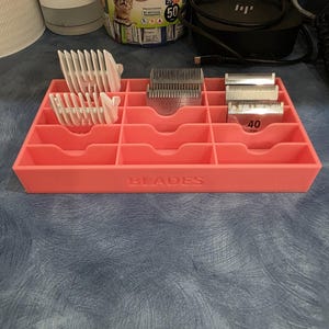 May include: A coral-coloured organiser labelled "BLADES" holds various grooming clipper blades and attachments. The organiser has multiple compartments to store different sizes and styles of blades. Some blades are labelled with the number "40".