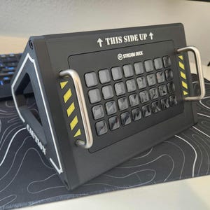 May include: A black Stream Deck device with multiple gray buttons, silver handles, and the text "THIS SIDE UP." The device has yellow diagonal stripes and the word "CAUTION" on the side. A keyboard and a black and white patterned mousepad are in the background.