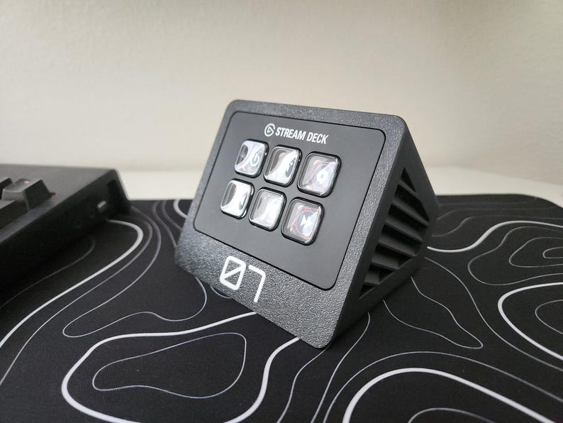 May include: A black Stream Deck with six illuminated buttons, the Stream Deck logo, and the number "07" in white. The device is angled and sits on a black and white patterned surface. A keyboard is partially visible in the background.