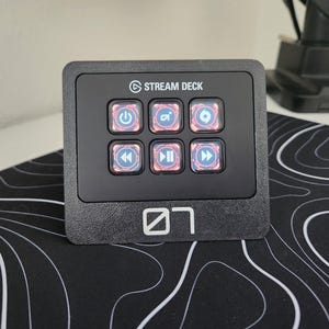 May include: Black Stream Deck with six illuminated buttons featuring various icons. The device has the "Stream Deck" logo and a white graphic. It sits on a black and white patterned surface.